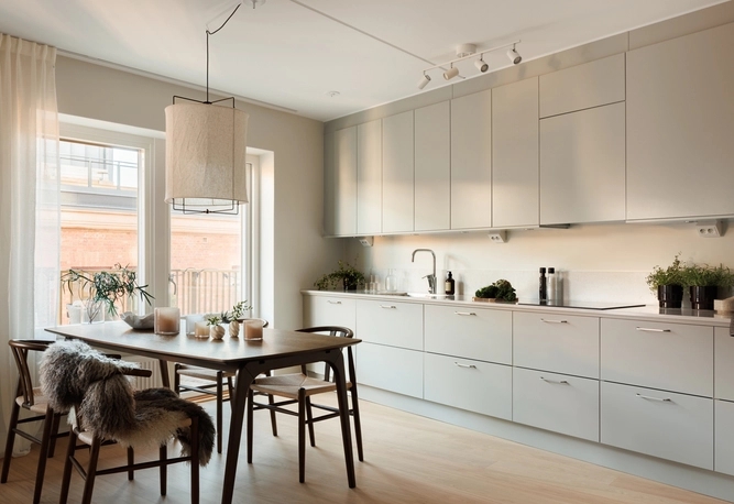 Ljust kök och matplats med moderna ljusgrå skåp, träbord med stolar och fällar, samt en taklampa. Minimalistisk stil.