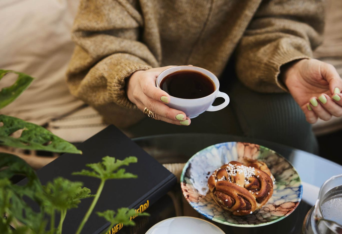 Person som håller en kopp kaffe och sitter nära ett bord med en kanelbulle på en färgglad tallrik