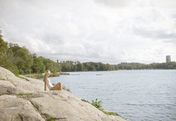 En kvinna sitter på en klippa vid en sjö, med träd och en hög byggnad i bakgrunden under en molnig himmel.