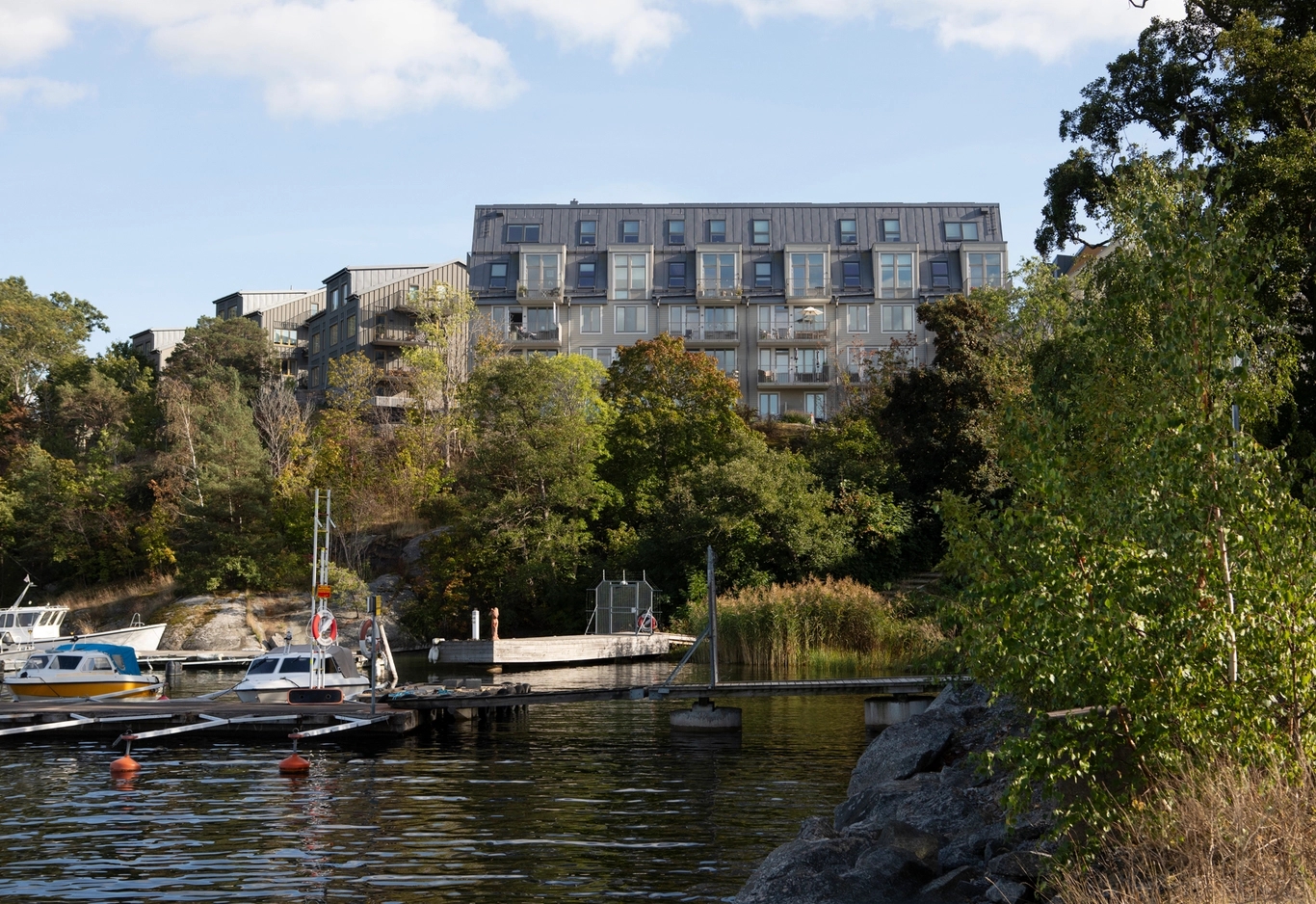 Brygga med båtar vid lugnt vatten, grönskande träd och ett flervåningshus i bakgrunden.