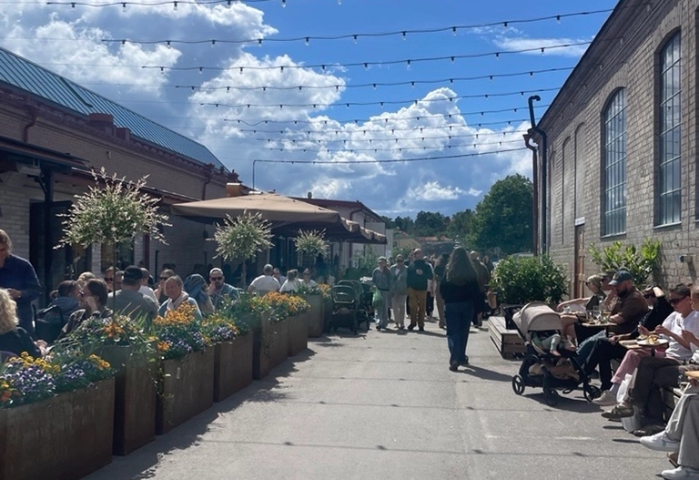 Livlig uteservering med många människor, bord, blommor och ljusslingor under blå himmel mellan tegelbyggnader.