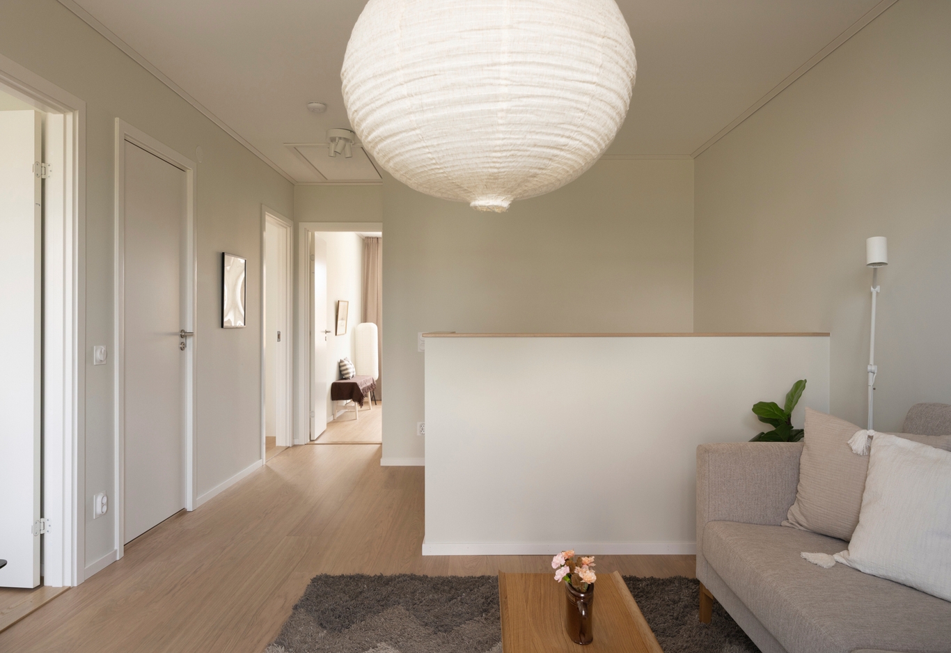 Ljust, modernt vardagsrum med trägolv, ljusgrå soffa, soffbord, matta och stor papperslampa. Hall till vänster.
