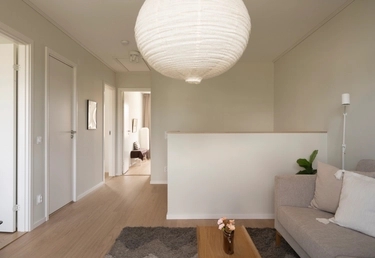Ljust, modernt vardagsrum med trägolv, ljusgrå soffa, soffbord, matta och stor papperslampa. Hall till vänster.