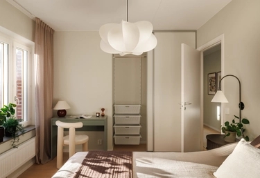 Ljust sovrum med modern taklampa, skrivbord vid fönstret, öppen garderob och säng med sängbord.