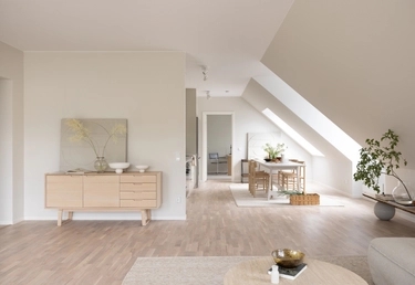 Ljust, minimalistiskt vardagsrum och matplats med ljusa trämöbler, vita väggar och ett stort snedställt fönster.