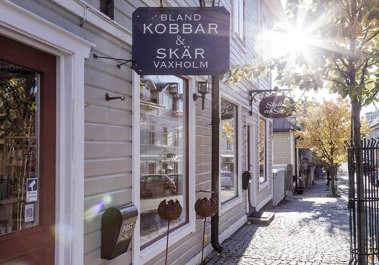 Gatuvy över en butik med en skylt med texten ”Bland Kobbar & Skär Vaxholm” på en solig dag med höstträd.