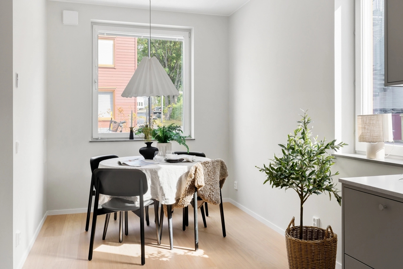 Ljust matrum med vitt bord, svarta stolar, vit veckad taklampa och krukväxt. Fönster med utsikt mot byggnad och grönska.