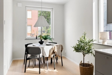 Ljust matrum med vitt bord, svarta stolar, vit veckad taklampa och krukväxt. Fönster med utsikt mot byggnad och grönska.