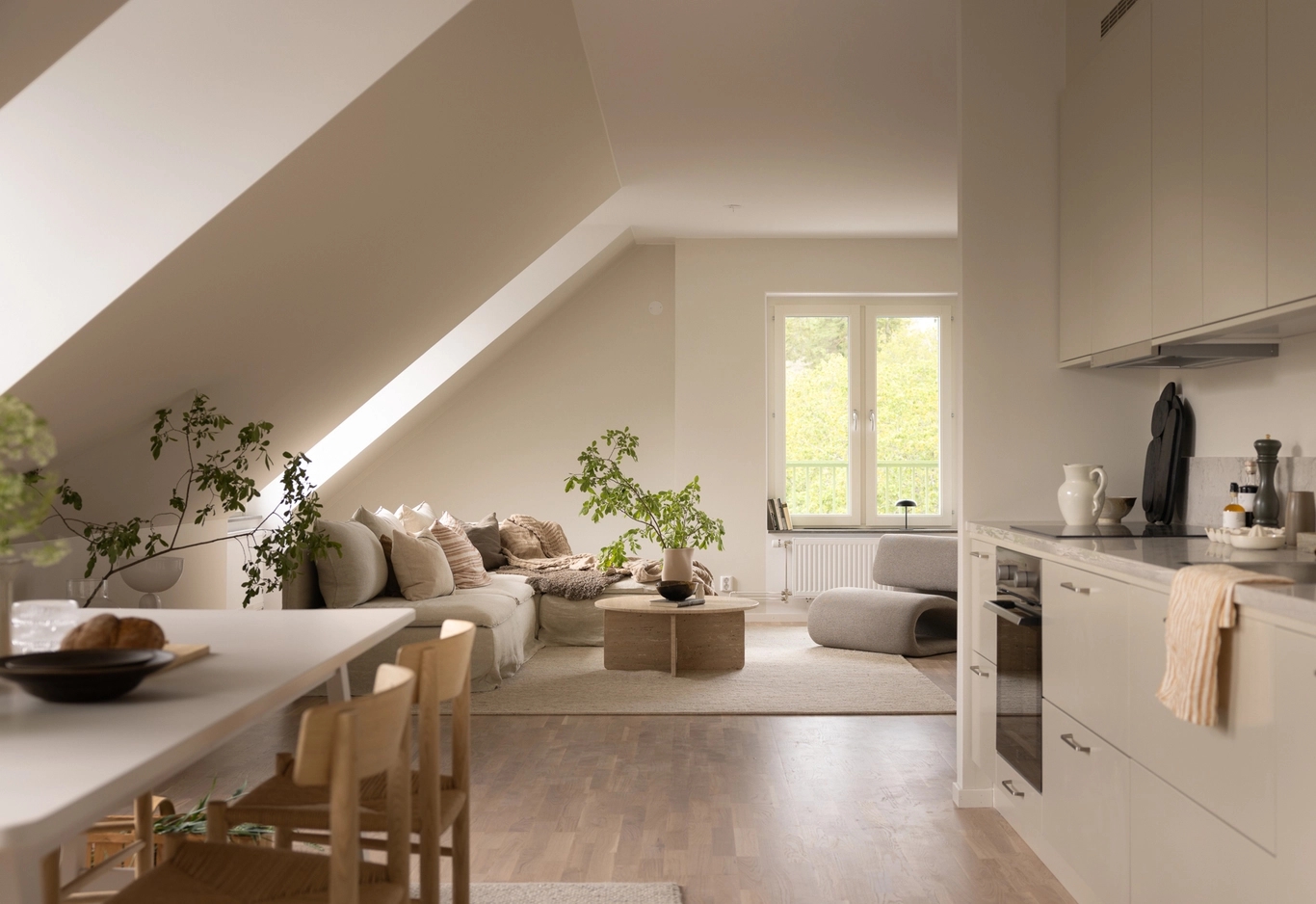 Modern vindslägenhet med neutral inredning, ett litet kök, matbord och en mysig vardagsrumsdel med växter vid fönstret.