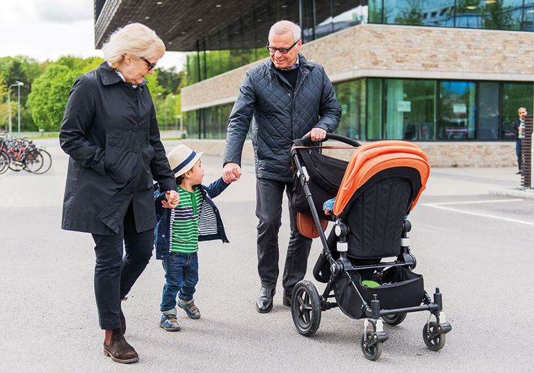 Två vuxna går med ett litet barn och en barnvagn utomhus på en asfalterad yta nära en modern byggnad.