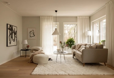 Neutral målat vardagsrum med beige soffa, runt soffbord, stora fönster och abstrakt konst på väggen.