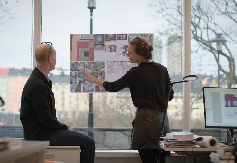 Två kollegor diskuterar designplaner som visas på en tavla i ett modernt kontor med stora fönster.