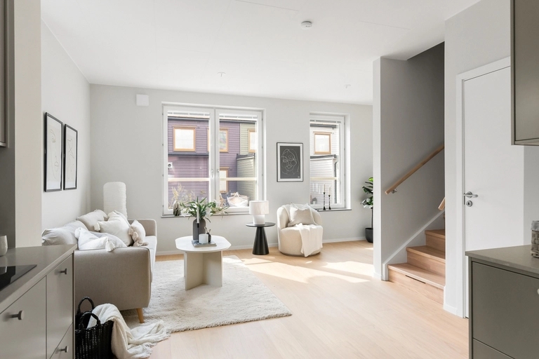 Ljust öppet vardagsrum med beige soffa, vit matta och ljust trägolv. En trappa med trästeg syns till höger.