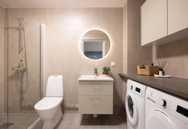 Modernt badrum med en rund belyst spegel, toalett, dusch, handfat och tvättmaskiner mot beige kaklade väggar.