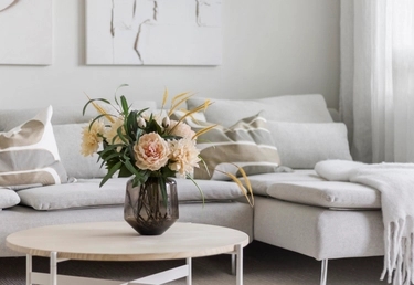 En beige soffa med mönstrade kuddar och schäslong vid ett runt bord med en vas med blommor i ett neutralt tonat vardagsrum.
