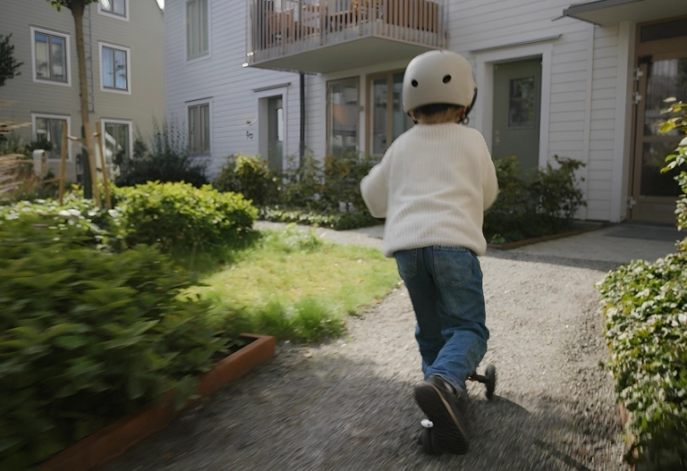 Ett barn med vit hjälm och tröja kör sparkcykel på en stig framför ett bostadshus.
