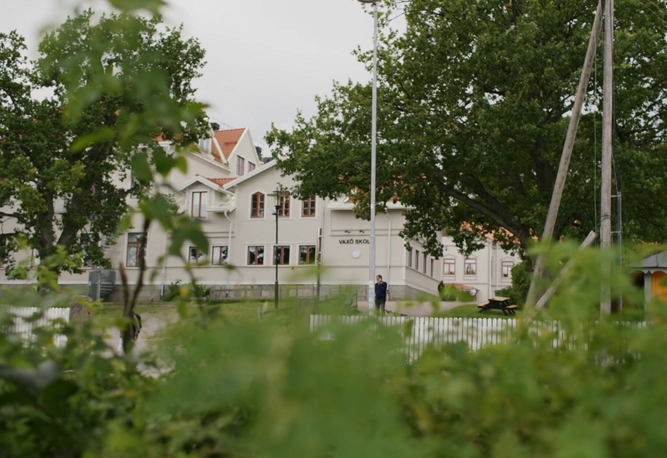 Stort vitt hus med röda tak syns delvis bakom gröna bladverk och träd en molnig dag.