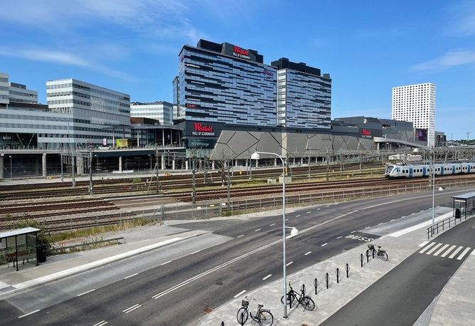 Tåg på spår i en stadsmiljö med moderna höghus, Mall of Scandinavia och en väg med cyklar i förgrunden.