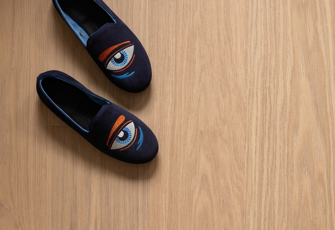 Ett par marinblå slip-on-skor med stiliserade ögonmönster på ett ljust trägolv.