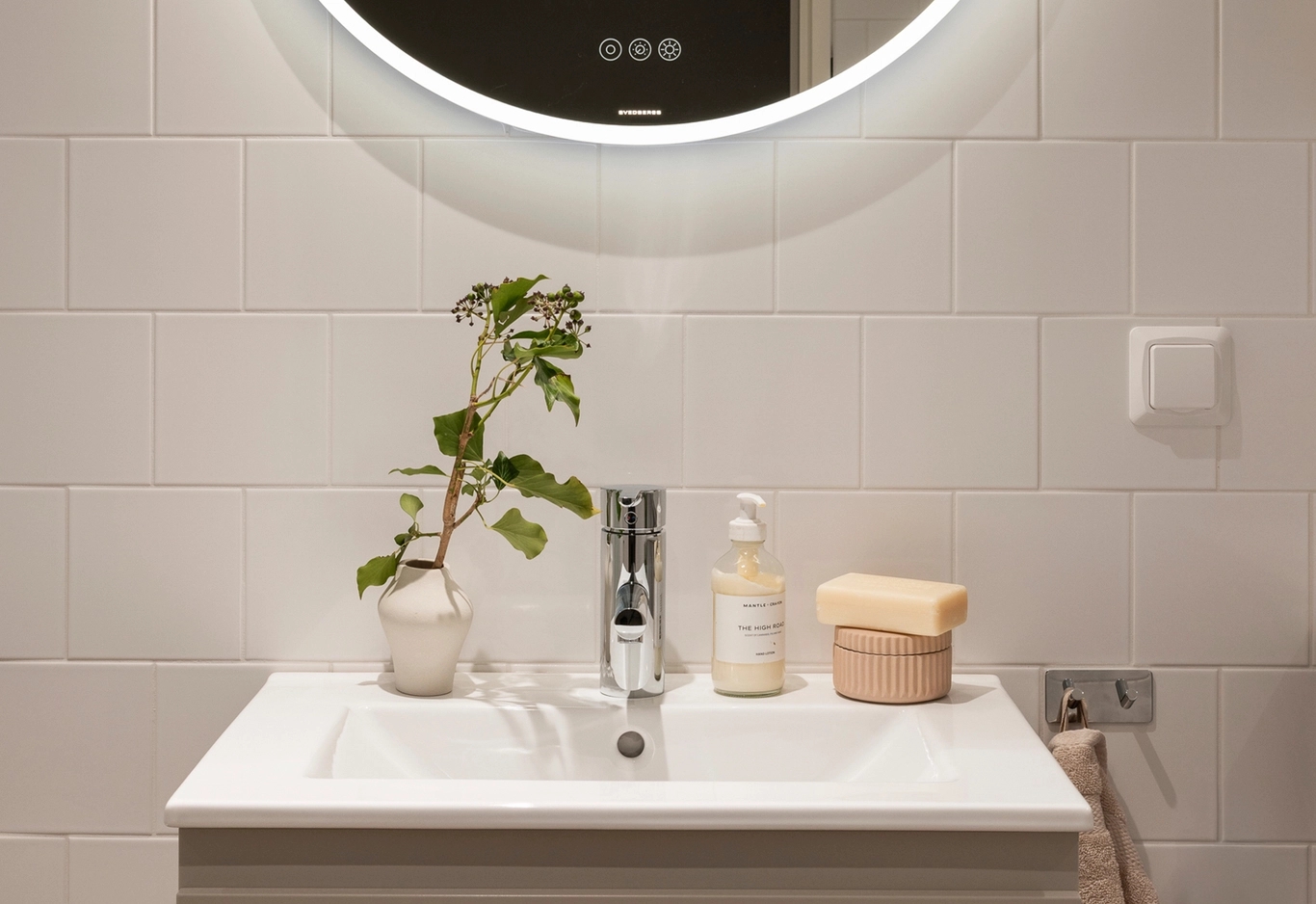 Ett handfat med en liten växt, tvålpump och tvål under en rund, belyst spegel på en kaklad vägg.