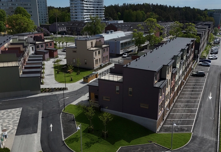 Flygbild över modernt bostadsområde med mörkbruna lägenhetshus, grönområden, gator och parkerade bilar under en ljus himmel.