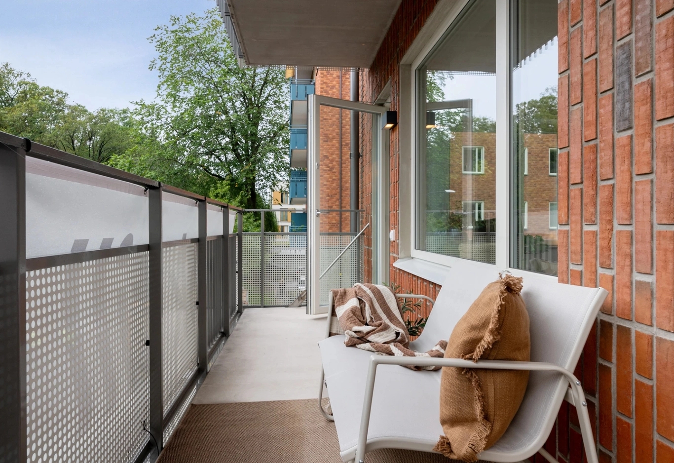 En balkong med en vit bänk, bruna kuddar, en filt och ett metallräcke med utsikt över träd och närliggande byggnader.