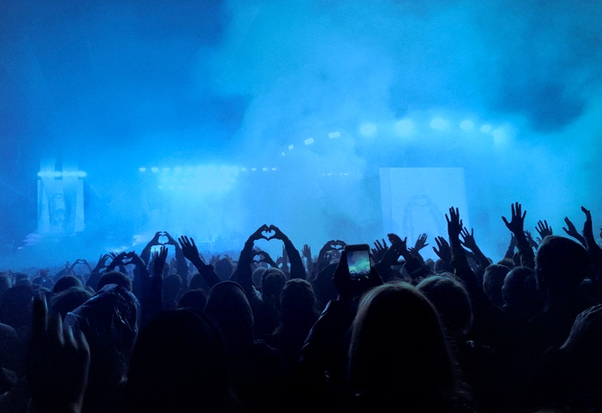 En publik på en konsert lyfter händer och telefoner i luften, omgiven av blå scenljus och rök.