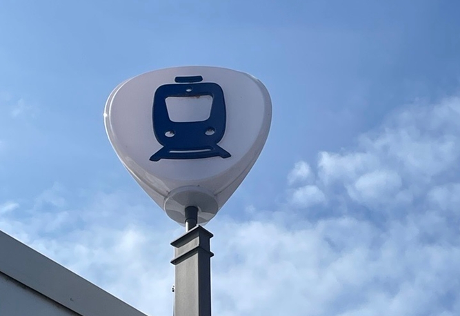 Vit upplyst skylt med blå pendeltågsikon på en stolpe mot en blå himmel med vita moln.