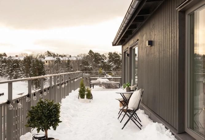 En snötäckt takterrass med ett litet bord, två stolar, krukväxter och utsikt över träd och hus i bakgrunden.