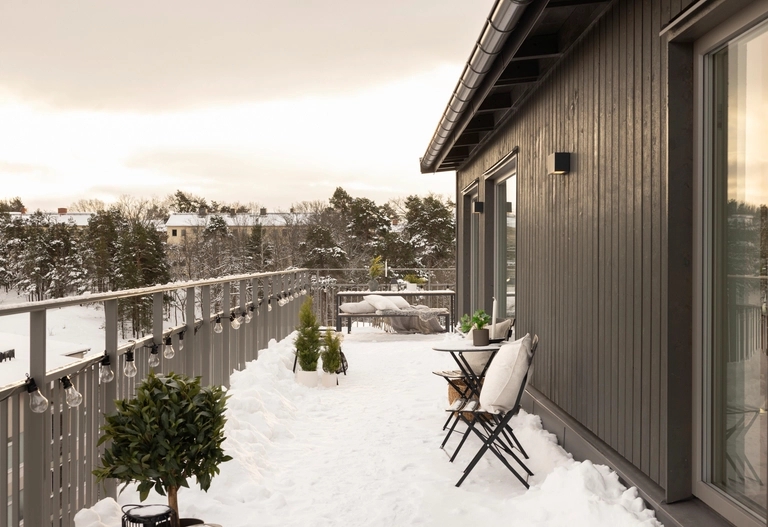 En snötäckt takterrass med ett litet bord, två stolar, krukväxter och utsikt över träd och hus i bakgrunden.