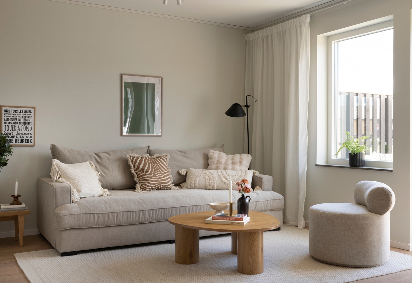 Modernt vardagsrum med beige soffa, runt soffbord i trä och fåtölj vid stort fönster med vita gardiner. Neutral inredning.