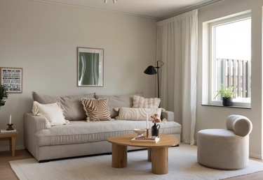 Modernt vardagsrum med beige soffa, runt soffbord i trä och fåtölj vid stort fönster med vita gardiner. Neutral inredning.