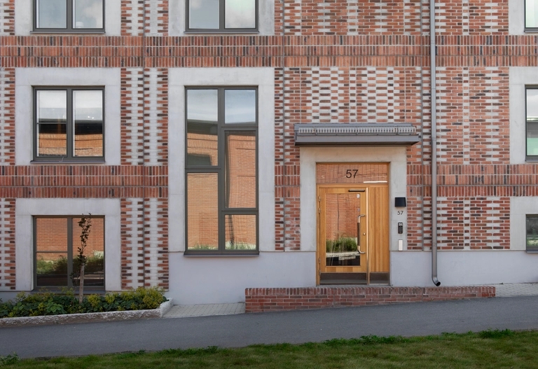 Rödbrun tegelfasad med mönstrade tegelstenar, mörka fönster och en ljus trädörr märkt 53. Gräsyta i förgrunden.