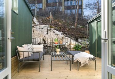 Litet utomhusdäck med en metallsoffa, kuddar, ett soffbord, krukväxter och en filt. Området är omgivet av ett räcke och omgivet av stenar och träd.