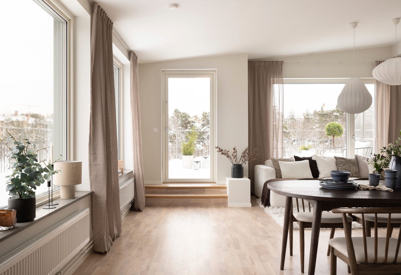 Ett ljust vardagsrum och matplats med stora fönster, ljusa trägolv och neutrala möbler, med utsikt över en snöig terrass.