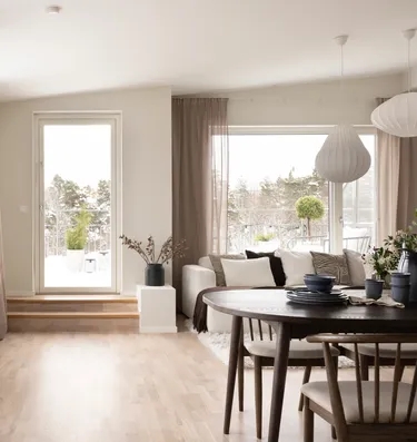 Ett ljust vardagsrum och matplats med stora fönster, ljusa trägolv och neutrala möbler, med utsikt över en snöig terrass.