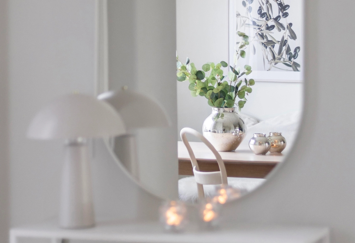 En vit lampa, ljus och bord reflekteras i en spegel som visar en vas med gröna grenar och ett inramat konstverk.