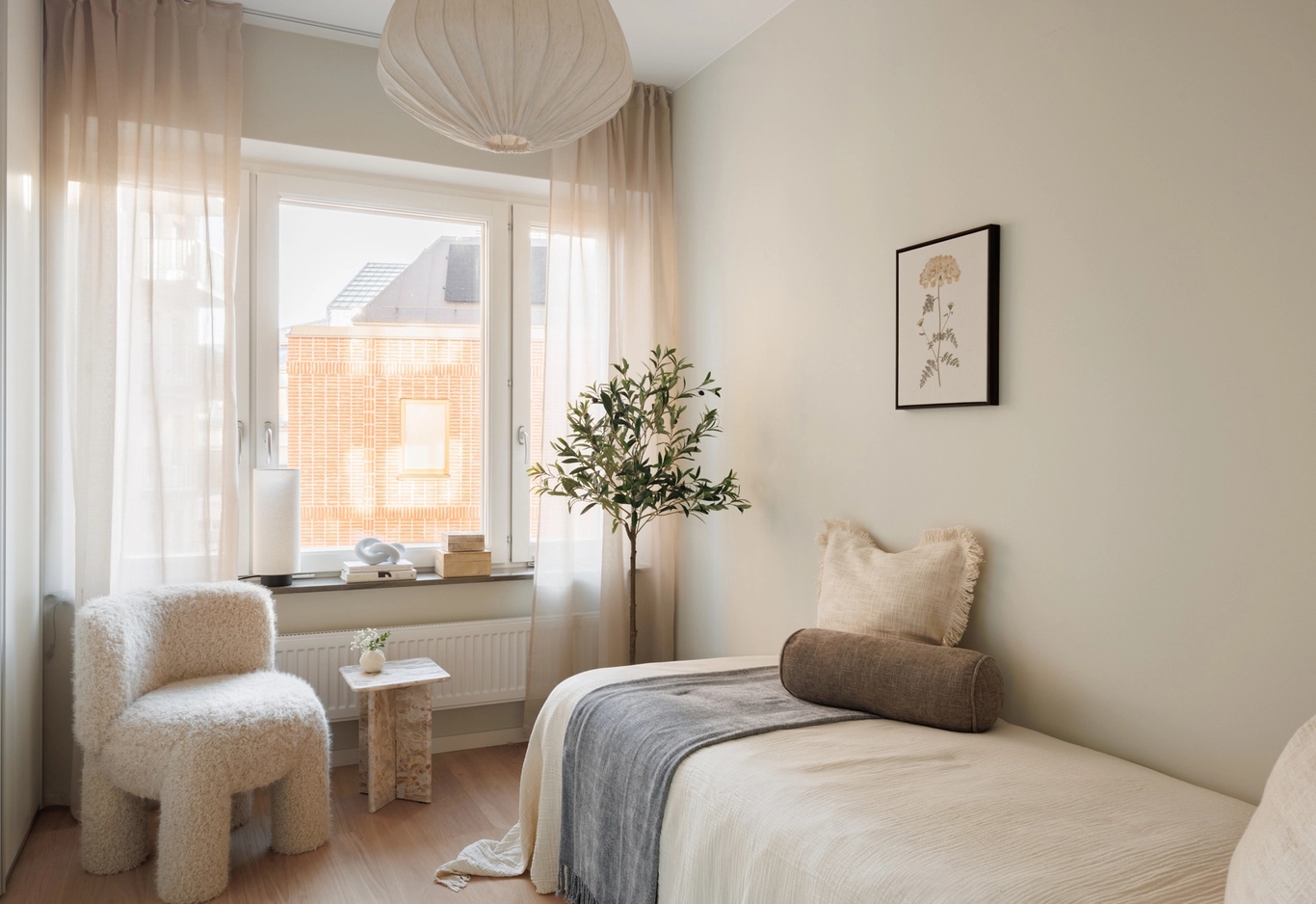 Sovrum med enkelsäng, neutrala färger, fönster med skira gardiner, luddig stol, krukväxt och botanisk poster.