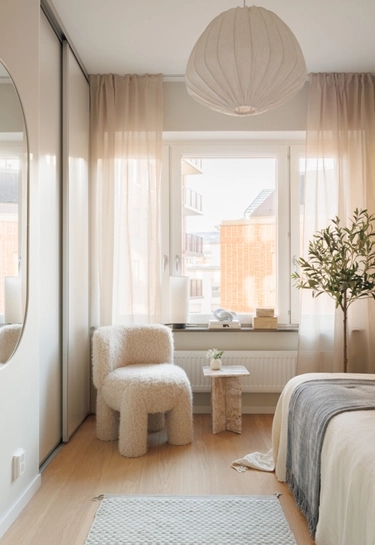 Ljust sovrum med gräddvit fluffig fåtölj, stort fönster, säng, en växt och modern taklampa.