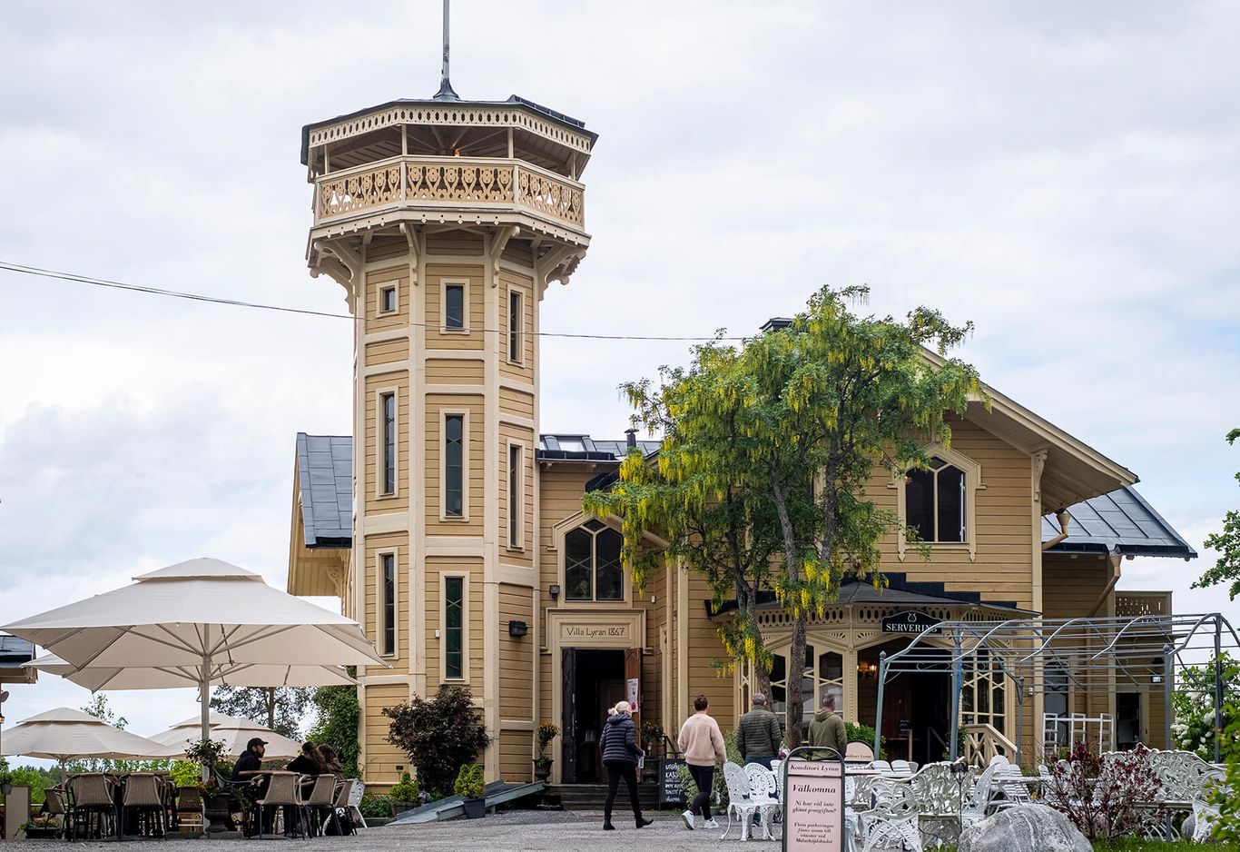 Gul träbyggnad med ett torn, uteservering med cafébord, konditori Lyran