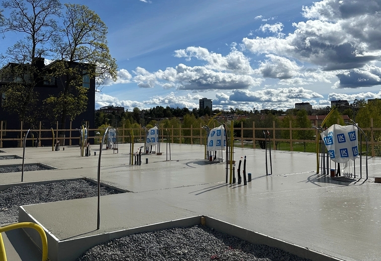 En byggarbetsplats med nygjutna betongplattor, utskjutande rör och synliga byggnader i bakgrunden.