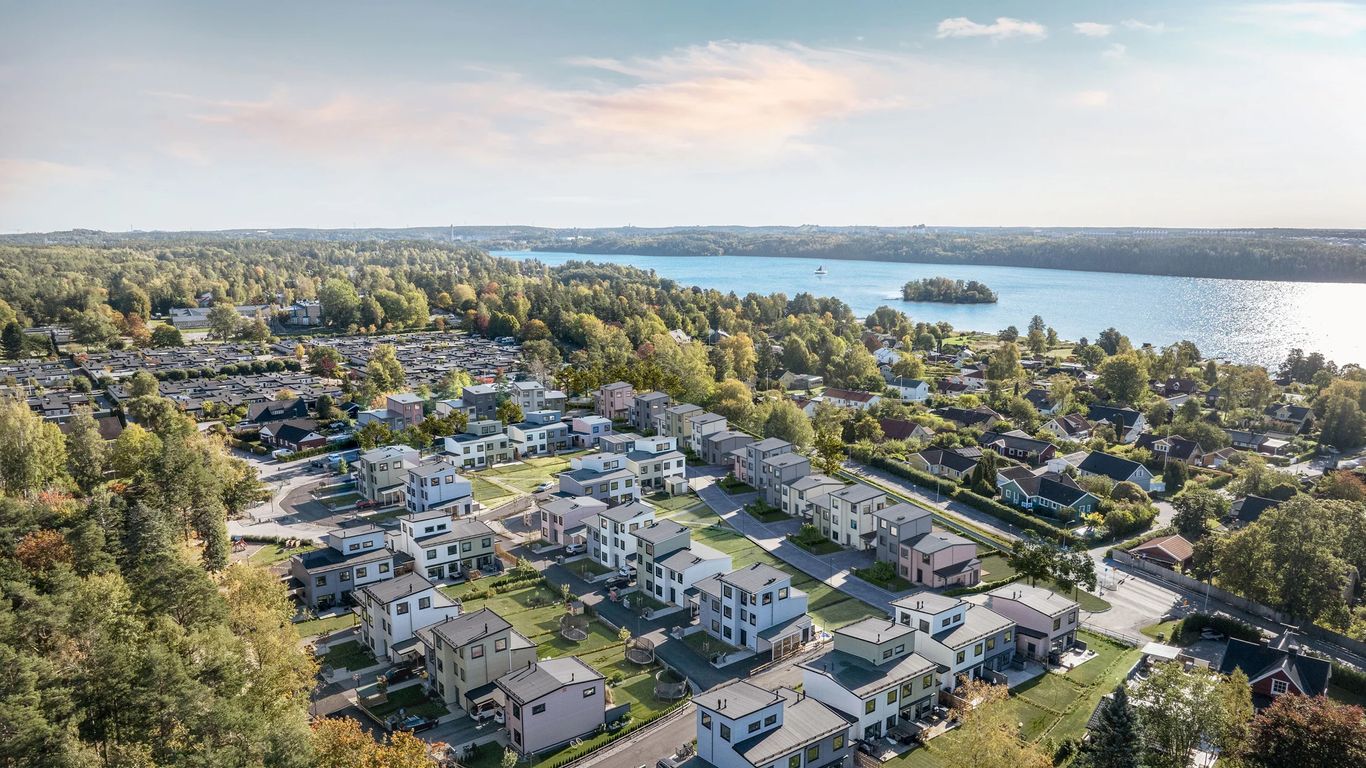 Flygfoto över ett bostadsområde med nybyggda och äldre hus nära en sjö och omgiven av träd.