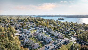 Flygfoto över ett bostadsområde med nybyggda och äldre hus nära en sjö och omgiven av träd.