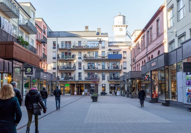Gågata kantad av butiker och lägenheter, människor som går och ett vitt torn syns i bakgrunden.