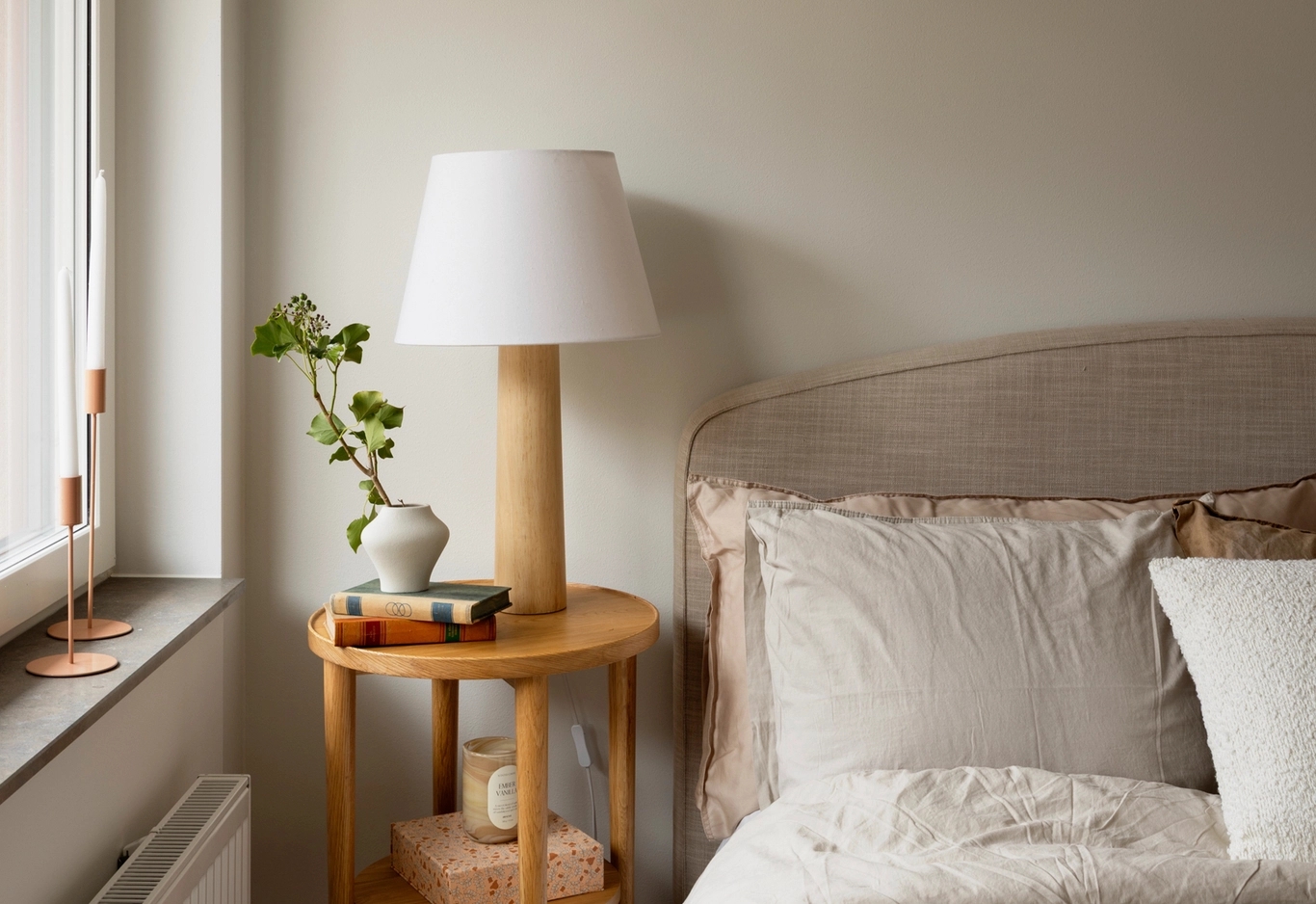Ett nattduksbord med en lampa, böcker, en vas med grönska, bredvid en snyggt bäddad säng med neutrala sängkläder.