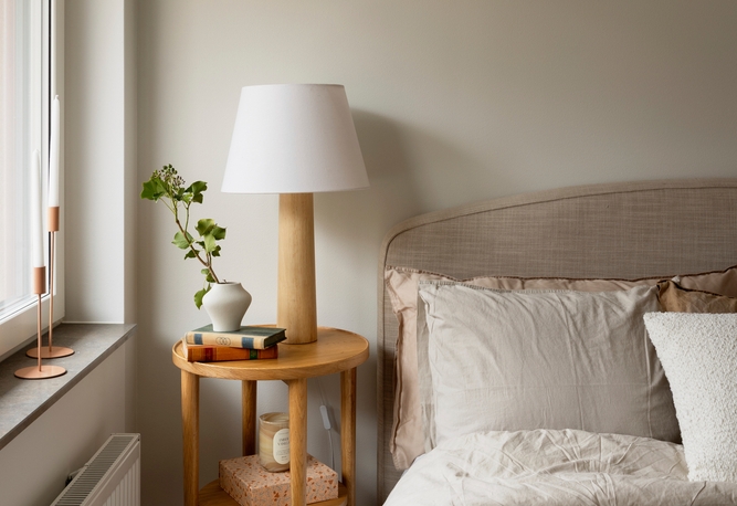 Ett nattduksbord med en lampa, böcker, en vas med grönska, bredvid en snyggt bäddad säng med neutrala sängkläder.