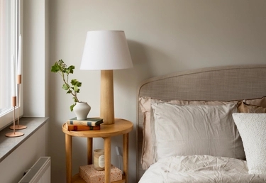 Ett nattduksbord med en lampa, böcker, en vas med grönska, bredvid en snyggt bäddad säng med neutrala sängkläder.