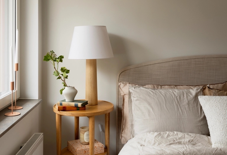 Ett nattduksbord med en lampa, böcker, en vas med grönska, bredvid en snyggt bäddad säng med neutrala sängkläder.