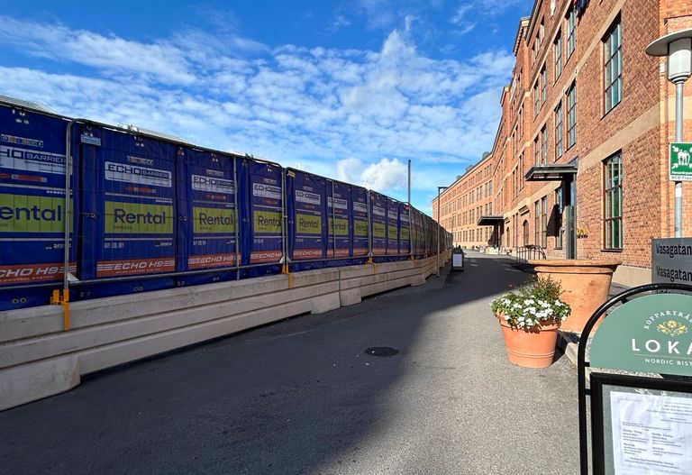Byggarbetsplats med blå barriär och tegelbyggnad till höger. En blomkruka och en skylt syns i förgrunden.