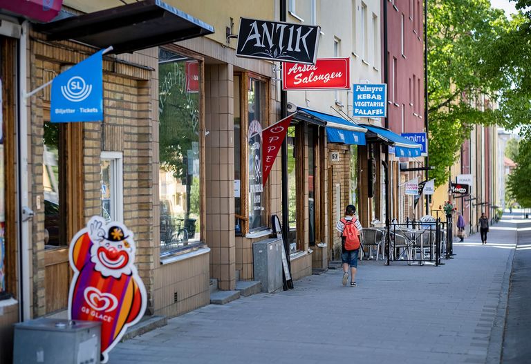 En fotgängare går förbi olika butiker och restauranger längs en solig trottoar kantad av skyltar och uteserveringar.